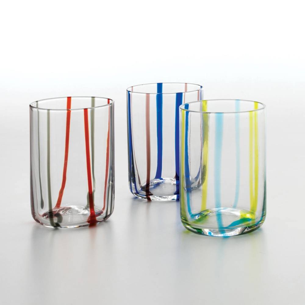 Tre bicchieri trasparenti in vetro con strisce verticali di colore rosso e grigio, blu e marrone, giallo e azzurro, su fondo bianco.