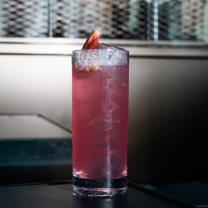 Hoog glas met transparante roze drank, ijs en citrusplakje op donker oppervlak met wazige donkere achtergrond