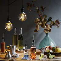 Bouteilles de liquides colorés en verre avec verseurs en métal sur table en bois, ampoules allumées, fruits et vase en céramique bleu clair avec