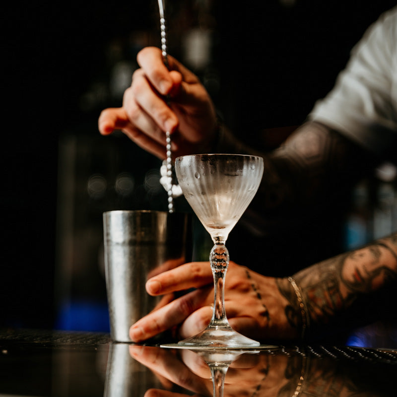 Glas met condensdruppels op reflecterend blad, metalen cocktail lamp en getatoeëerde hand die een drink roert.