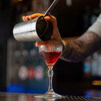Getatoeëerde hand schenkt rode vloeistof uit metalen shaker in transparante glazen kelk in wazige bar