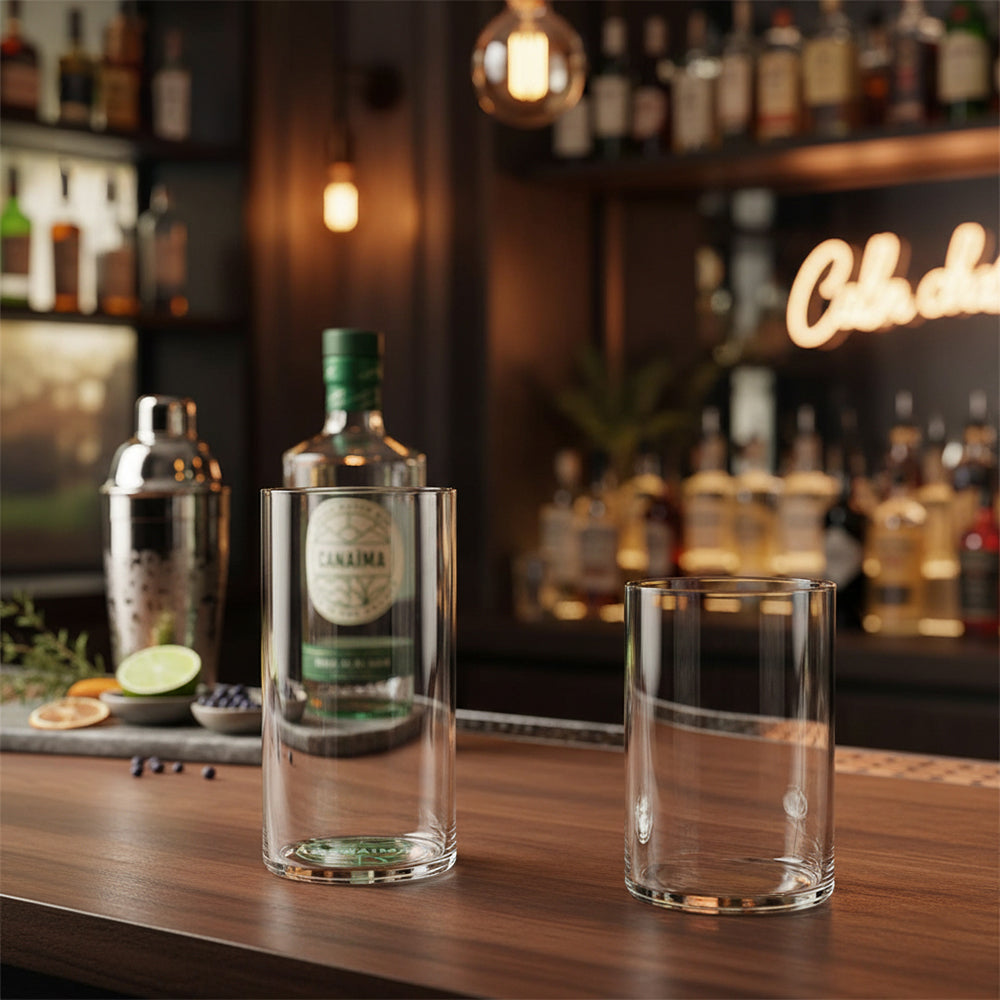 Glass bottle with green "Canaima" label and two transparent glasses on wooden bar counter with fruit and shaker in bar with warm lights