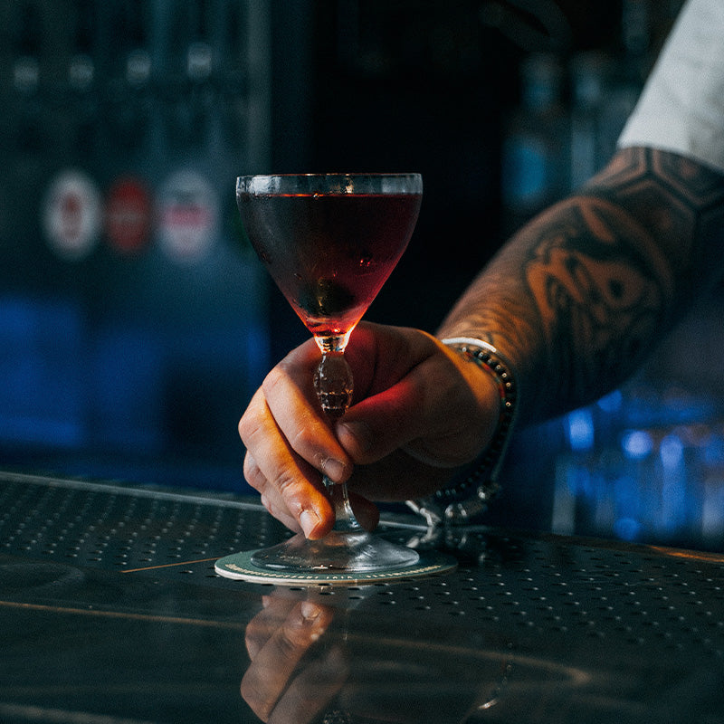 Hand met armbanden en tatoeages houdt cocktailglas met donkerrode vloeistof op reflecterend metalen blad