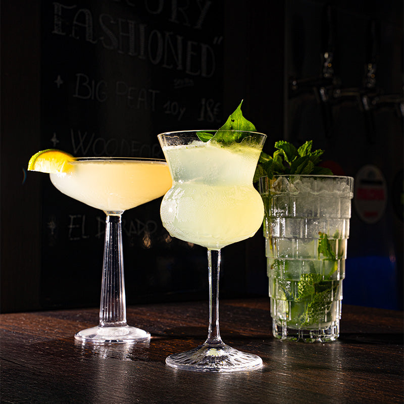 Tre cocktail diversi con ghiaccio, foglie di menta e fette di limone, su tavolo di legno scuro in ambiente bar.