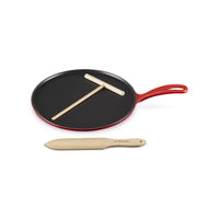 Padella rotonda rossa con manico, superficie nera, e due utensili in legno, uno a forma di T e uno piatto, in un ambiente neutro.
