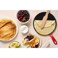 Crespelle dorate, frutti di bosco in ciotola, panna in tazza, limoni, spatola di legno su fondo chiaro.