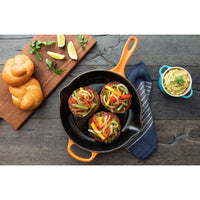 Trois hamburgers avec légumes colorés dans une poêle noire. À côté, petits pains dorés, citron vert et sauce verte en bol. Ambiance rustique.