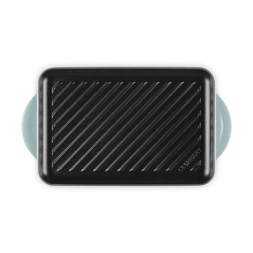 Teglia rettangolare nera con manici turchesi, superficie rigata, materiale apparentemente metallico o ceramico, sfondo bianco.