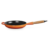 Poêle avec fond noir et bord orange, manche en bois, surface lisse, posée sur fond blanc.