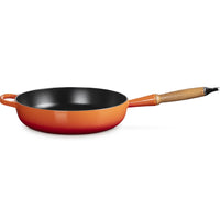 Padella rotonda con bordo arancione sfumato verso il rosso, interno nero lucido, manico in legno e metallo.