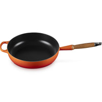 Padella rotonda con manico in legno, esterno arancione e interno nero. Superficie liscia e lucida, senza alimenti visibili.