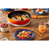 Padella arancione con toast dorati, piatti blu con frutta e dessert, bicchiere di succo e fragole in ciotola su tavolo di legno.