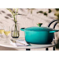 Aqua green casserole with golden lid and handle on a marble table, flanked by wine glasses and cutlery.