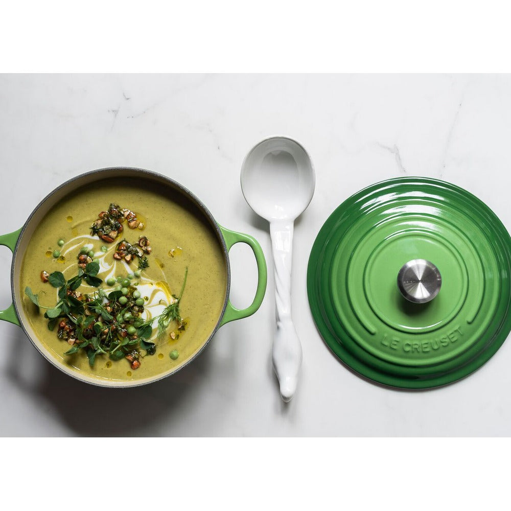 Green soup in green metal pot, decorated with herbs and crunchy elements; white spoon on the side and green lid on top.