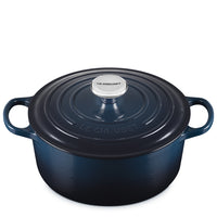 Dark blue oval enameled cast iron pot with lid, two side handles and metal knob with Le Creuset inscription, white background.