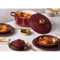 Red casserole with golden lid, matching plates, roast chicken with vegetables, light napkin and wine glasses.