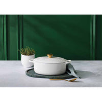 Casserole blanche avec poignée dorée sur un plateau vert, accompagnée d'un vase blanc avec herbes et d'une cuillère dorée. Fond vert.