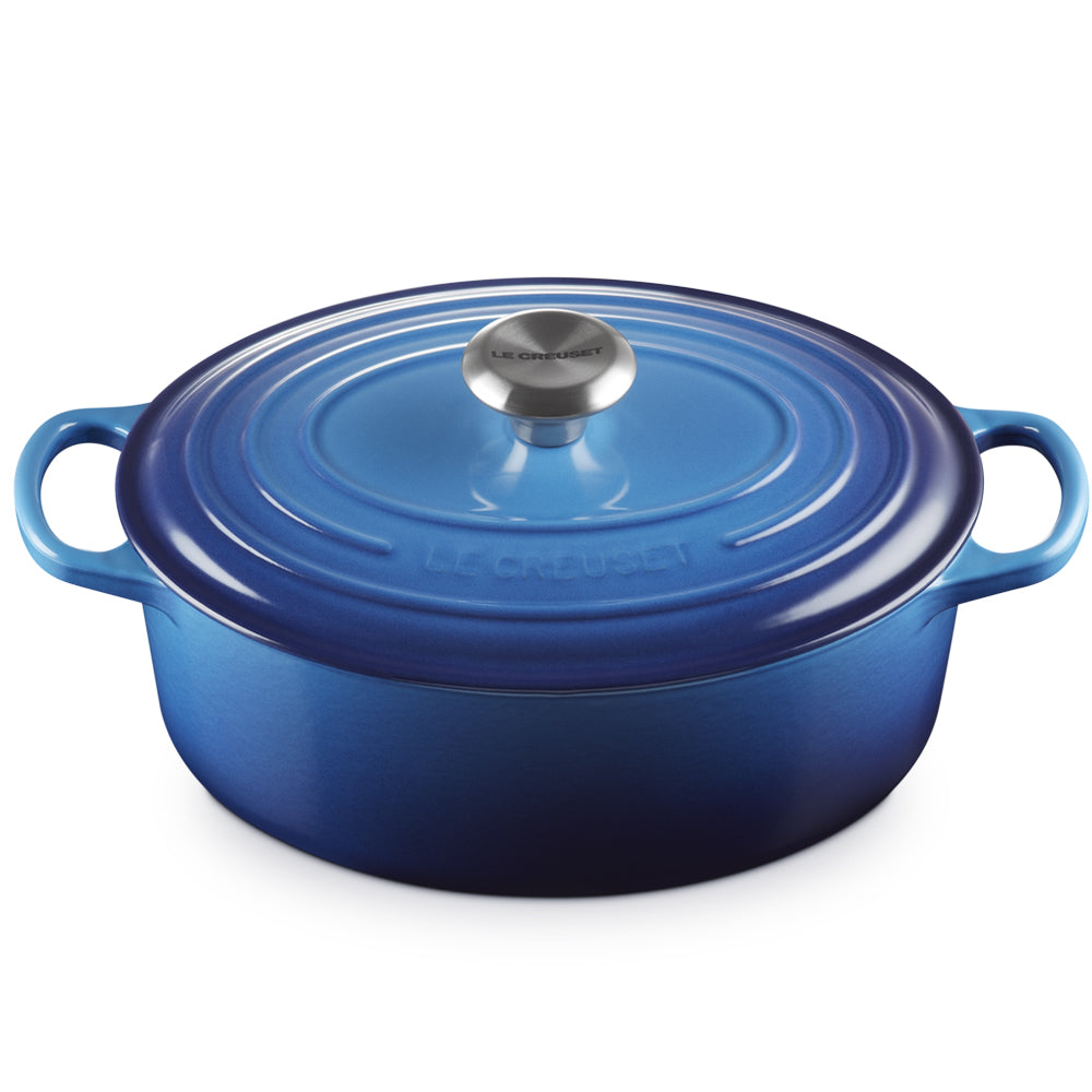 Casserole ovale bleu en fonte émaillée avec couvercle et deux anses, bouton métallique avec inscription Le Creuset, fond blanc.