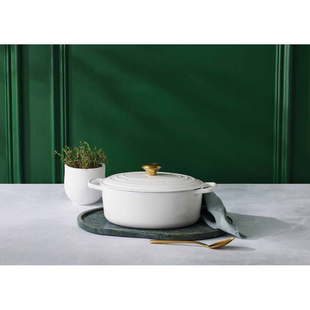 Witte braadpan met gouden deksel op groen dienblad, naast witte vaas met plant en gouden lepel. Groene achtergrond.