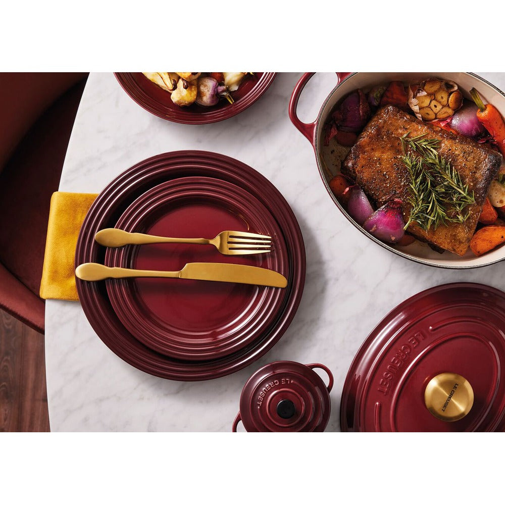 Gedekte tafel met bordeauxrode borden, gouden bestek, een gele servet en een pan met vlees en groenten. Elegante sfeer.