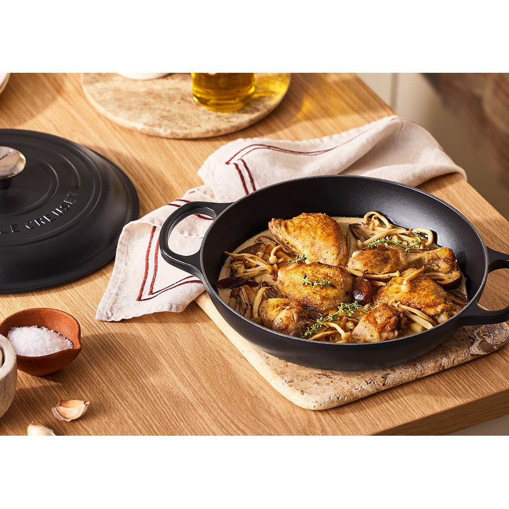 Pentola in ghisa nera con pollo dorato e funghi su un tagliere di legno. Sullo sfondo, olio d'oliva e tovagliolo bianco con strisce rosse.