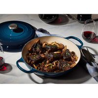 Pentola in ghisa smaltata blu con carne e funghi, guarnita con prezzemolo, e bicchiere di vino rosso sul tavolo.