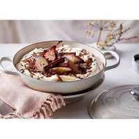 Ciotola bianca con dessert a base di panna, pere caramellate e scaglie di cioccolato, decorata con bastoncini di cannella, appoggiata su un piano