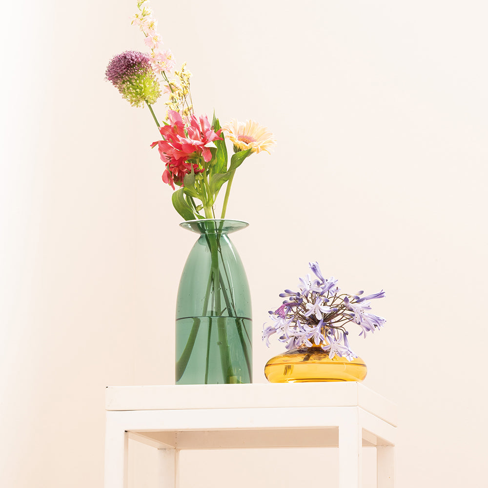 Green vase with colorful flowers and yellow vase with purple flowers, on a white table, on light background.