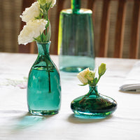 Two green glass vases with white flowers on a white table, a glass decorative element in the background.