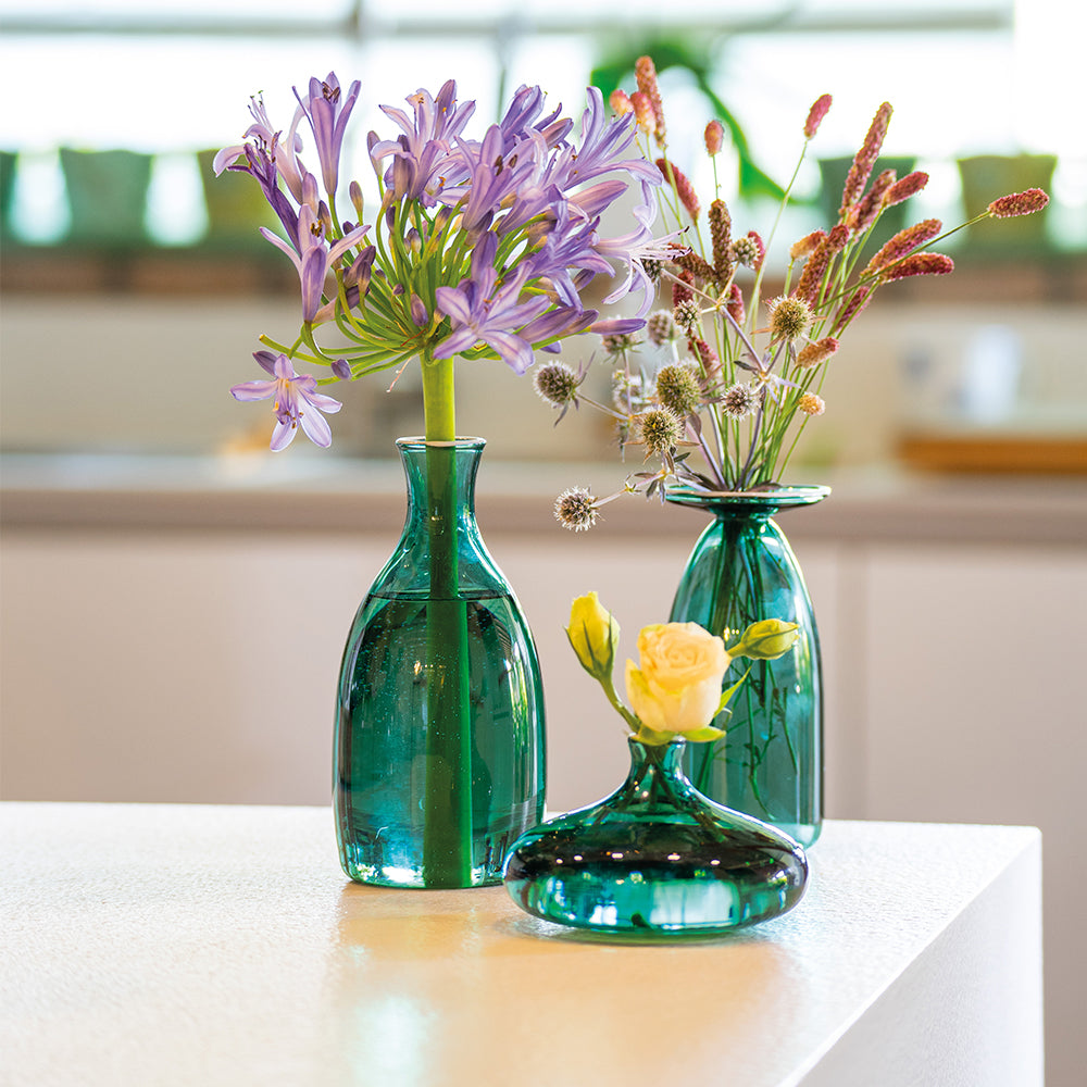 Trois vases verts en verre avec fleurs : un avec fleurs violettes, un avec épis secs et un avec une rose jaune. Fond lumineux.