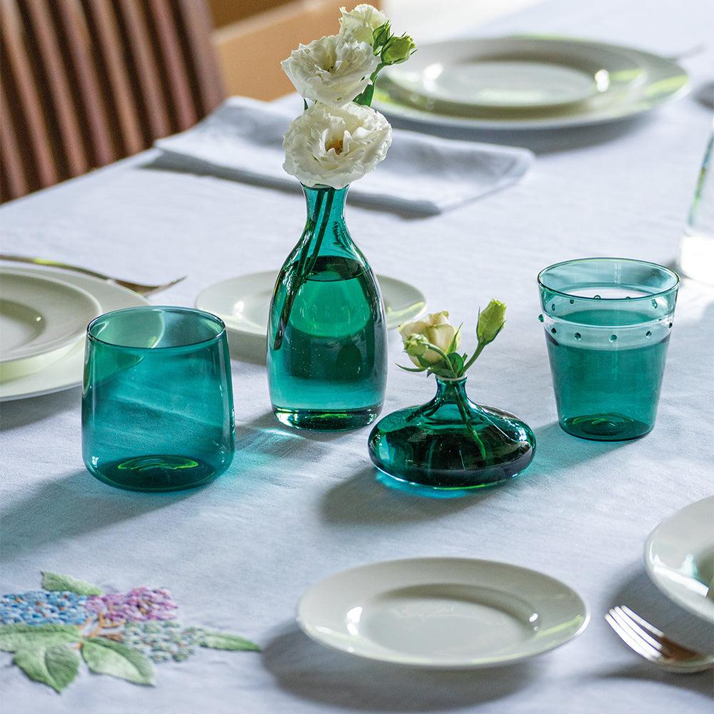 Vases en verre vert d'eau avec fleurs blanches, assiettes claires et nappe blanche à broderie florale. Cadre élégant et lumineux.