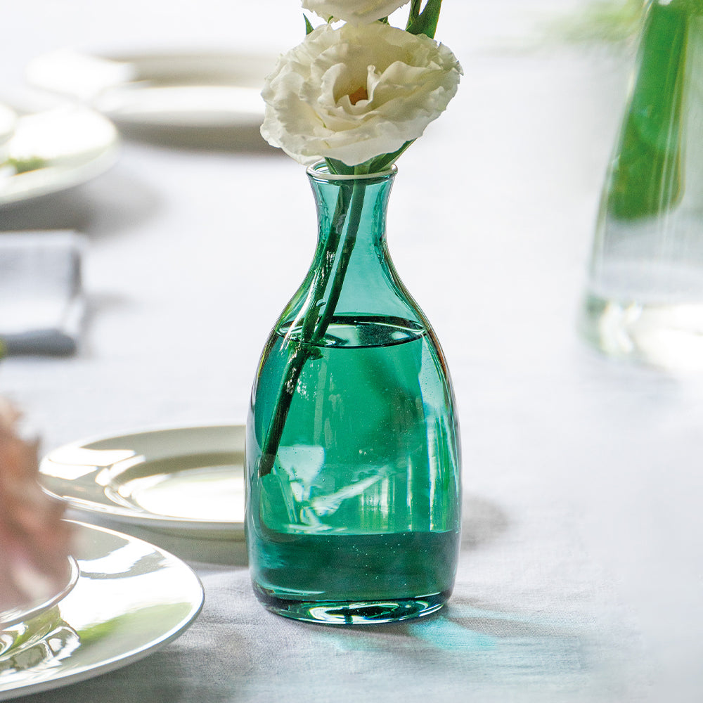 Green glass vase with white flowers, on a light tablecloth, surrounded by plates and another vase.