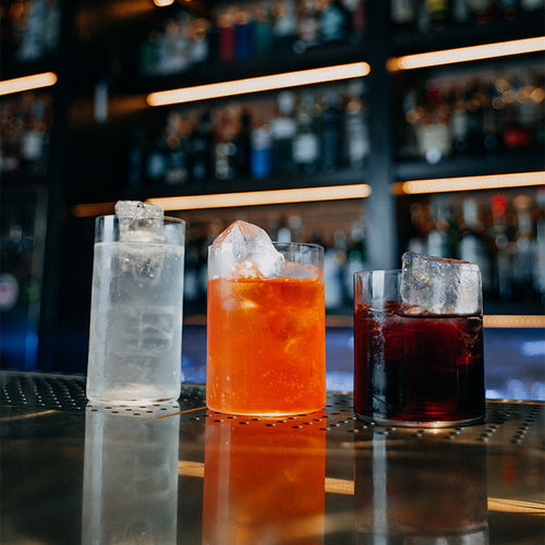 Tre bicchieri trasparenti con liquidi di colore bianco, arancione e rosso scuro con ghiaccio, su bancone metallico in bar.