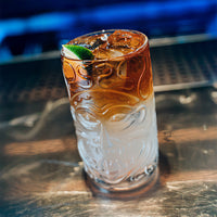 glass tumbler with stylized face pattern containing cocktail a strati marrone and transparent with ice and lime slice green