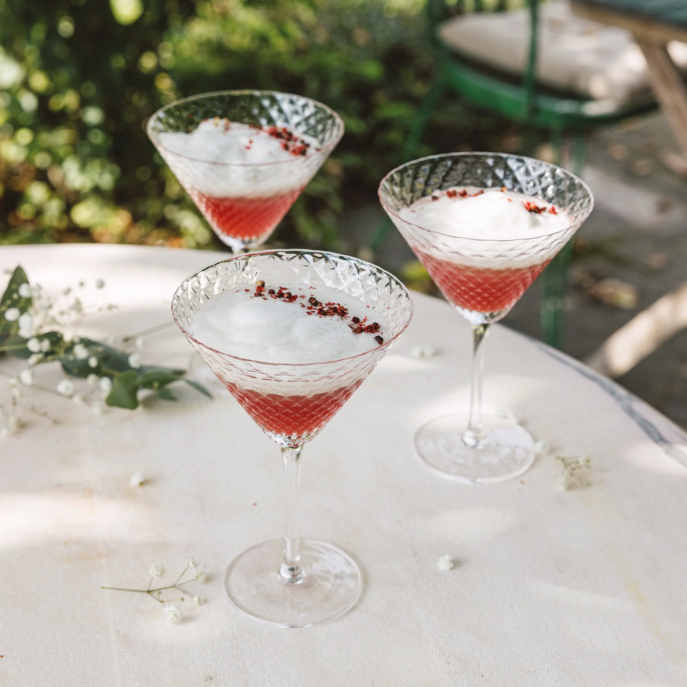 tre cocktail rosa con schiuma bianca e piccoli petali rossi in bicchieri di vetro inciso su tavolo chiaro all'aperto