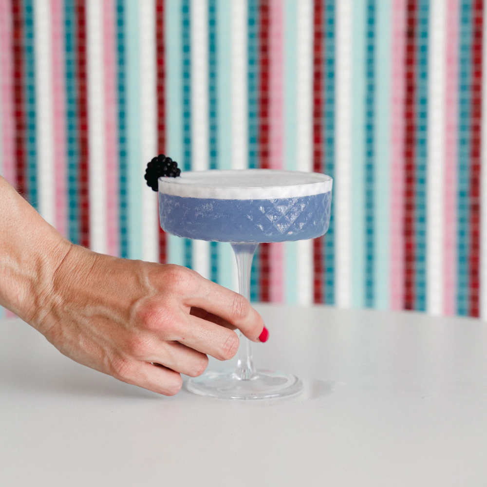 Main au vernis rose tenant un calice en verre avec cocktail violet à mousse blanche et mûre sur fond à rayures colorées
