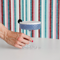 Main au vernis rose tenant un calice en verre avec cocktail violet à mousse blanche et mûre sur fond à rayures colorées