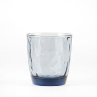 Transparent glass tumbler with wavy shape, light gray color and base blue scuro, on white background.