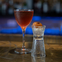 bicchiere da cocktail in vetro trasparente con liquido rosso e bicchierino decorato con cubetti di ghiaccio e frutto arancione su tavolo