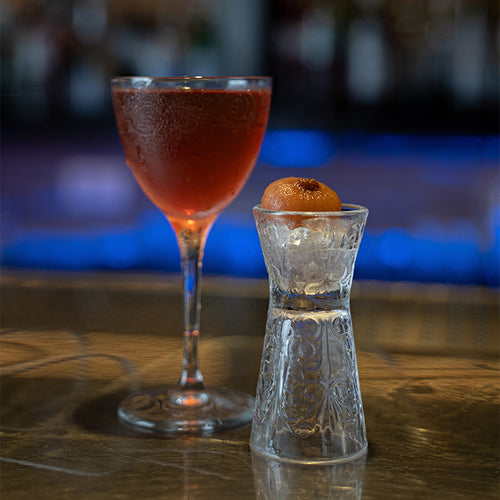 bicchiere da cocktail in vetro trasparente con liquido rosso e bicchierino decorato con cubetti di ghiaccio e frutto arancione su tavolo