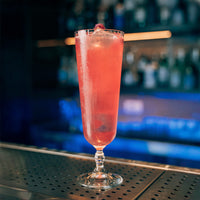 bicchiere di vetro alto con cocktail rosa e ghiaccio, decorato con lampone, su bancone metallico in bar sfocato sullo sfondo
