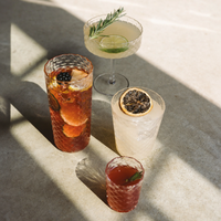 Cuatro vasos de vidrio facetado con cócteles coloridos, decorados con rodajas de fruta y ramitas, sobre superficie de cemento claro