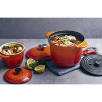 Deux casseroles en céramique orange avec couvercle, une plus grande avec soupe, tranches de citron et épices sur fond gris.