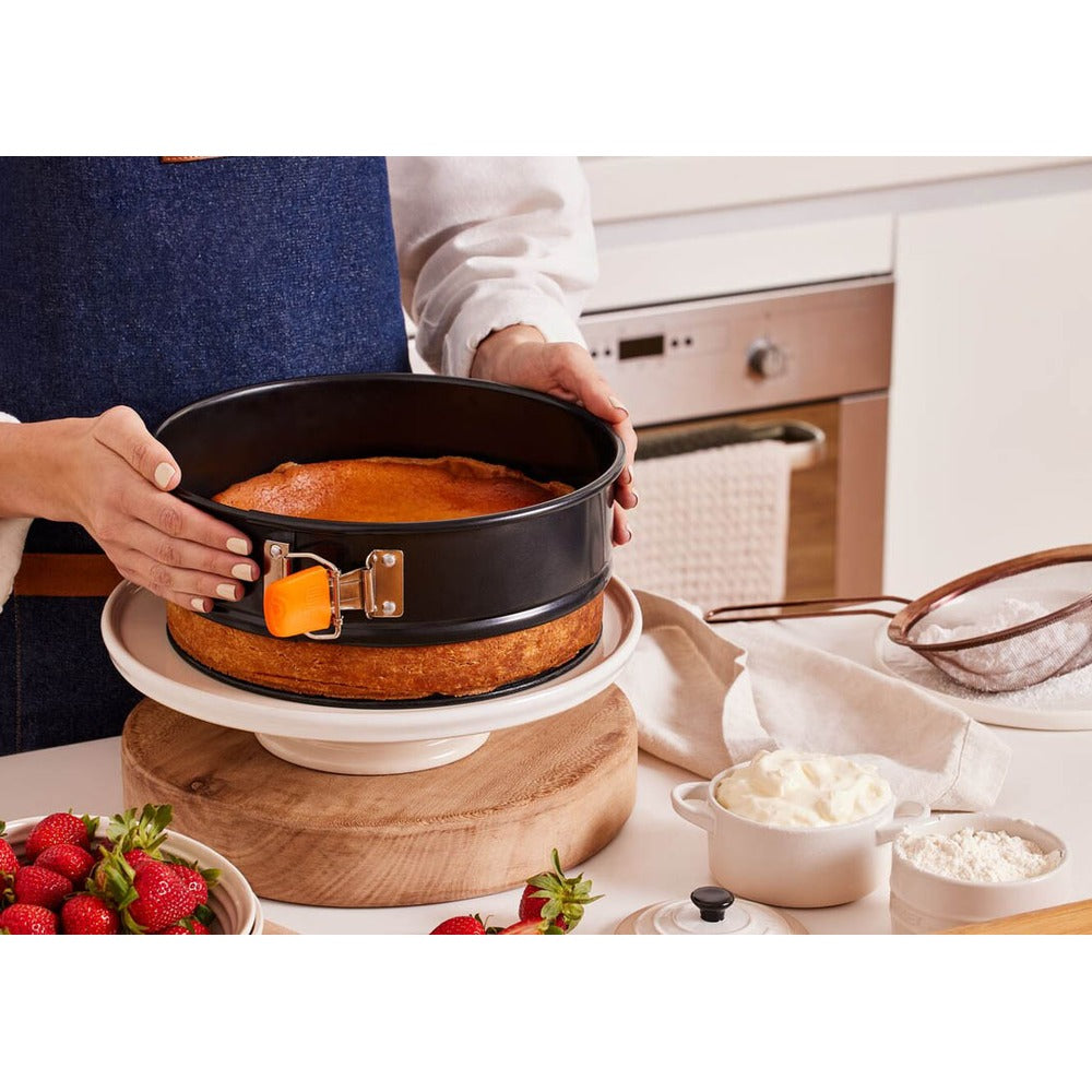 Freshly baked cake in a black round baking pan, placed on a white plate; strawberries and whipped cream visible on the table.