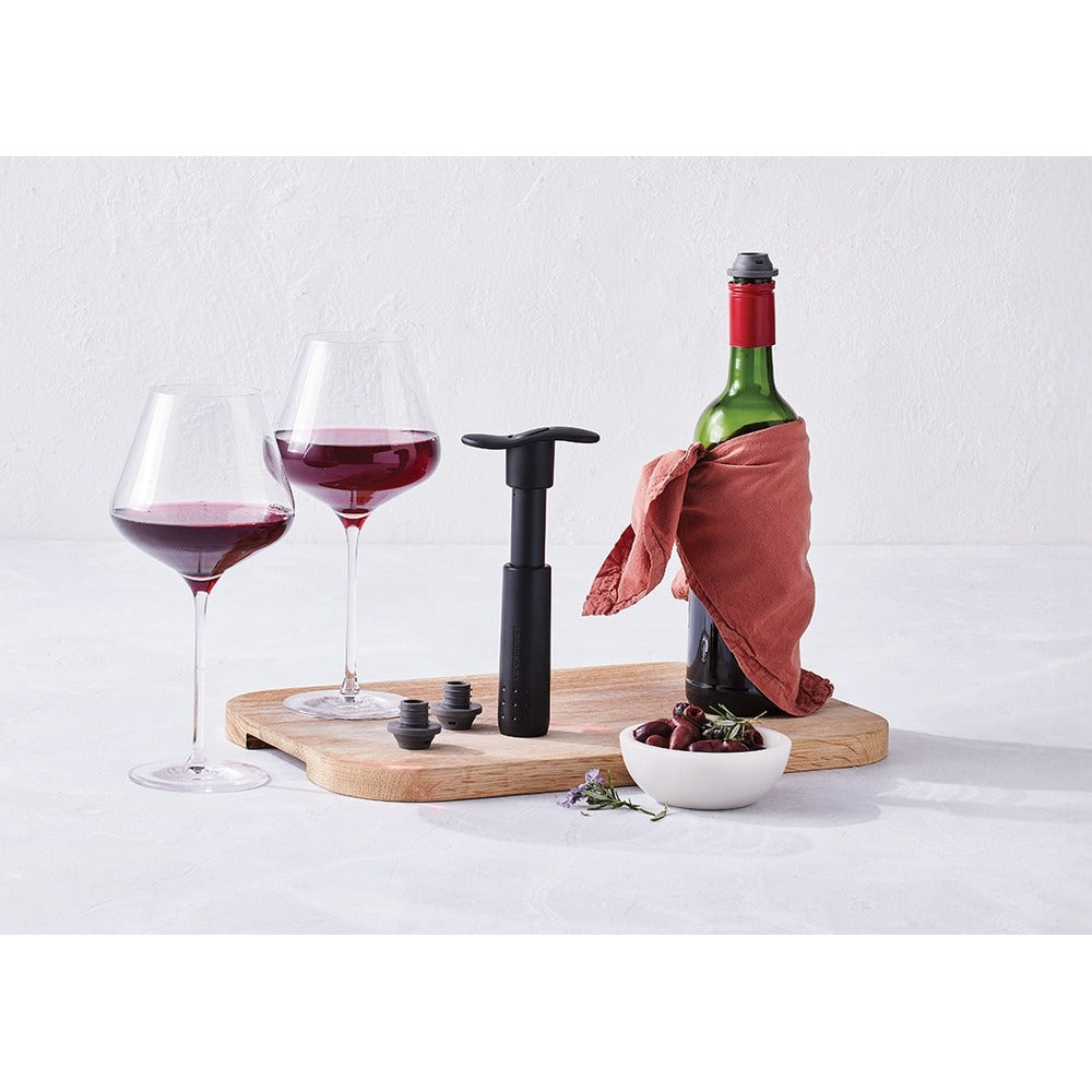 Due calici di vino rosso, una bottiglia con un panno rosso, un tappo nero e una ciotola con ciliegie su un tagliere di legno.