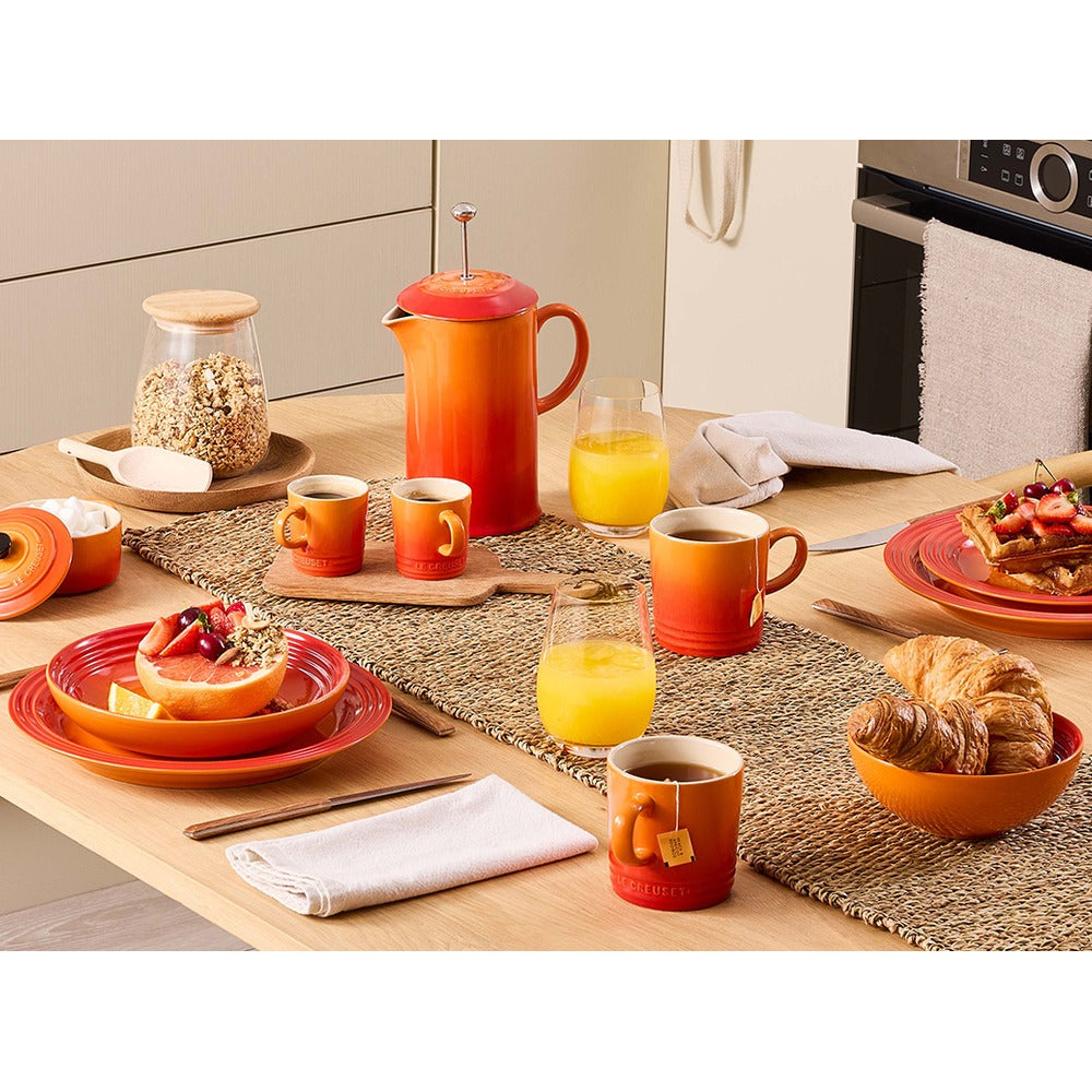 Tavola apparecchiata con piatti, tazze e una caffettiera arancione, accompagnati da frutta, croissant e succo d'arancia.