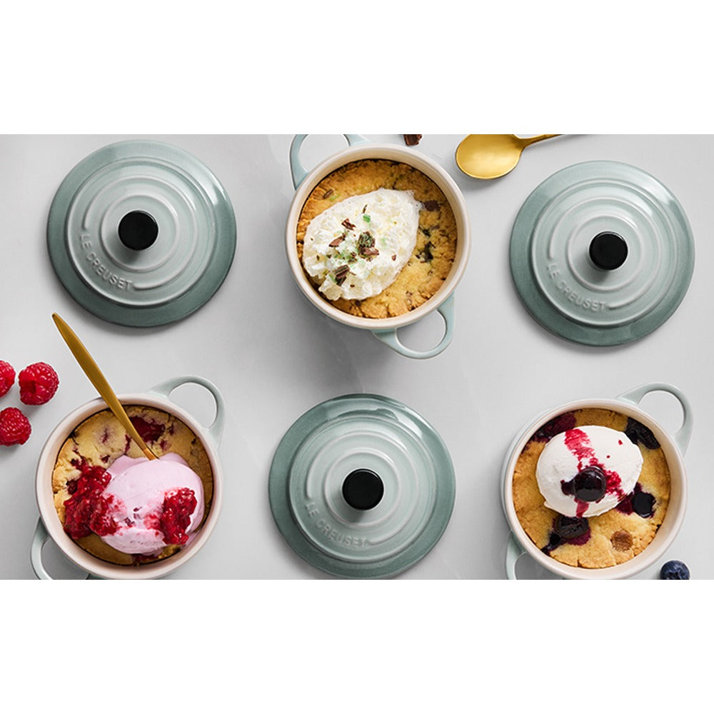 Beige and green ceramic bowls with desserts and ice cream, garnished with red berries, and golden spoon beside. Neutral setting.