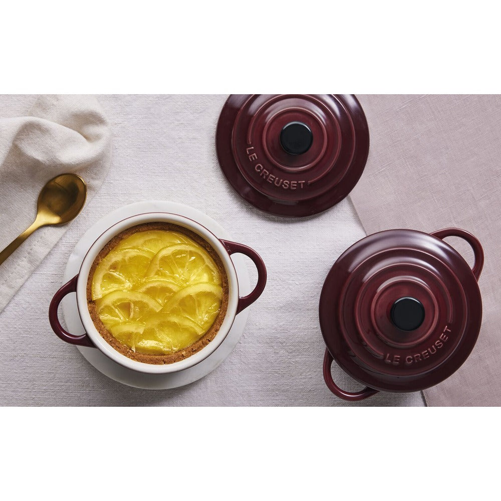 White cake pan with decorated lemon dessert, near two burgundy lids and a golden spoon on light fabric.