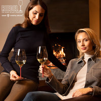 two women in autumn outfits hold white wine glasses with red stem in front of a lit fireplace in cozy modern setting