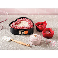 Tarta en forma de corazón decorada con frambuesas, espátula blanca con corazones rojos, cuencos rojos en forma de corazón y cacerola rosa. Mesa gris.